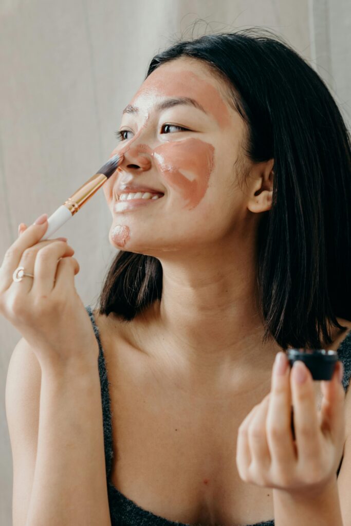 Smiling woman applies clay mask using a brush, focusing on skincare routine.