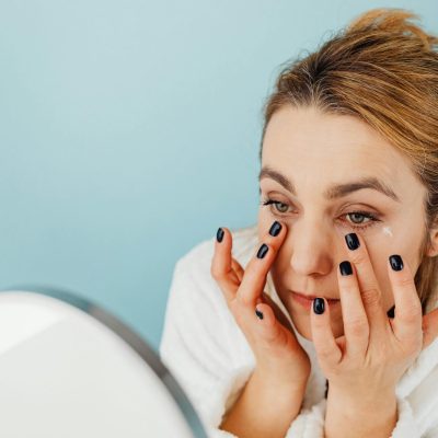 Woman applies eye cream on her under-eye area with black nails, wearing a white robe.